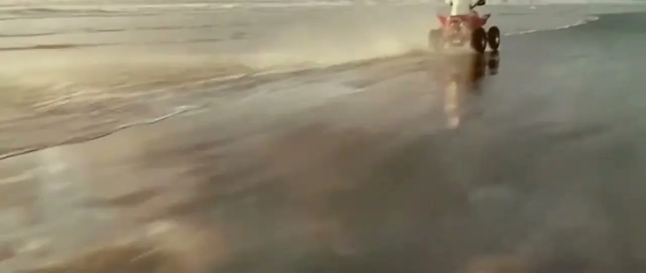 A person in white rides a red ATV along a wet beach at golden hour, leaving a trail of spray behind them as they speed across the sand.