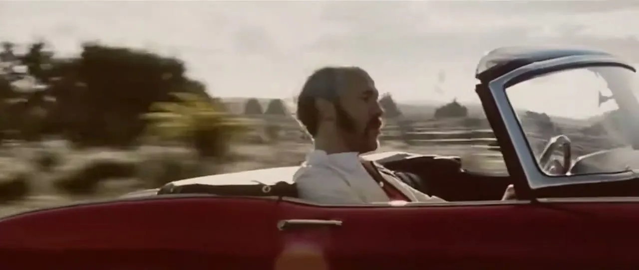 A man with a beard drives a red convertible car on a sunny day, with the landscape blurring past him.