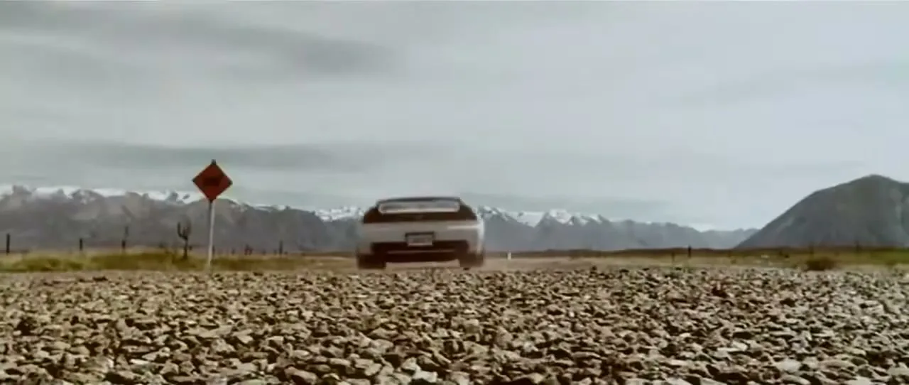 A white Honda car drives away from the camera on a gravel road, with snow-capped mountains in the background under a cloudy sky.
