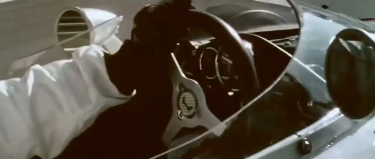 A person in a white racing suit and black gloves drives a vintage open-wheel race car on a track, with the camera focused on their hands on the steering wheel.