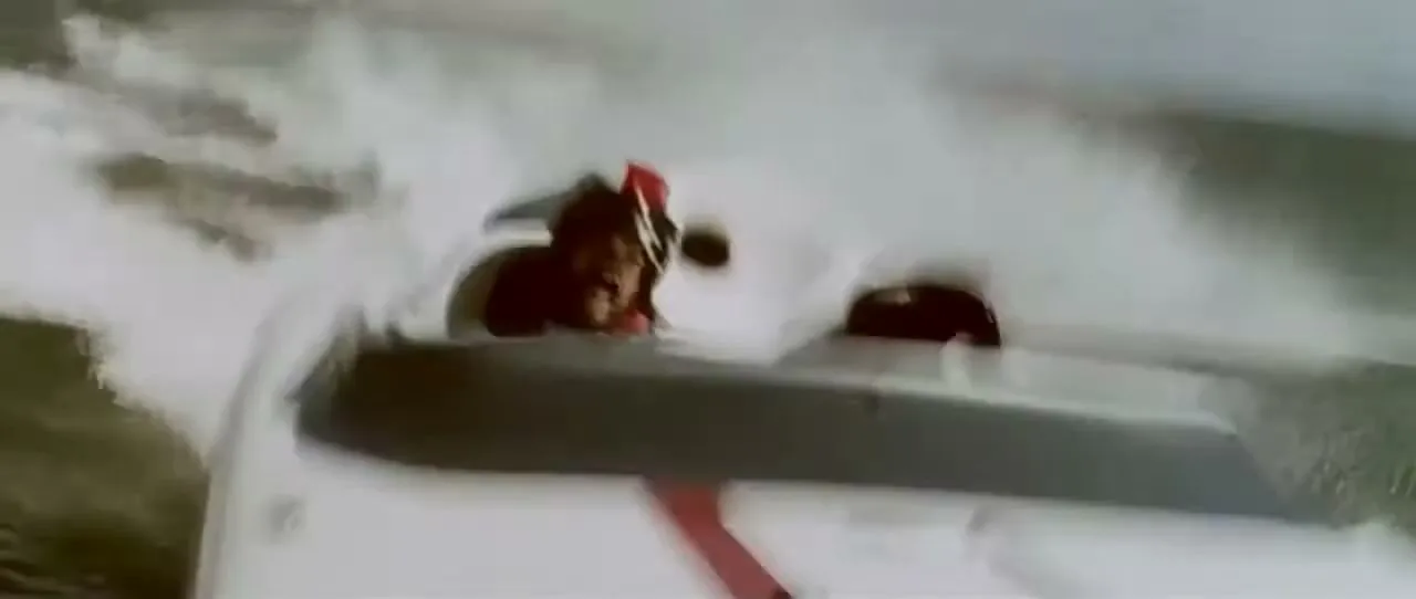 A man with a mustache and helmet drives a white and red speedboat, creating a large wake on the water.