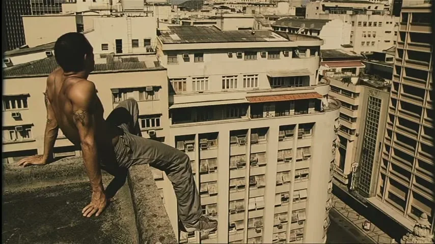 A shirtless man with tattoos sits on the edge of a tall building, looking out over a dense city landscape on a sunny day.