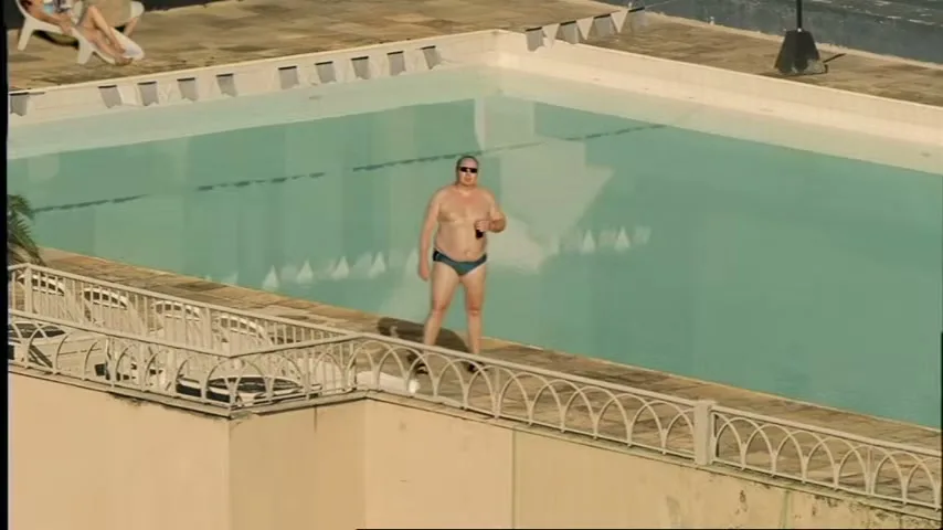 A bald, overweight man in a speedo and sunglasses stands by a rooftop pool, holding a black object.