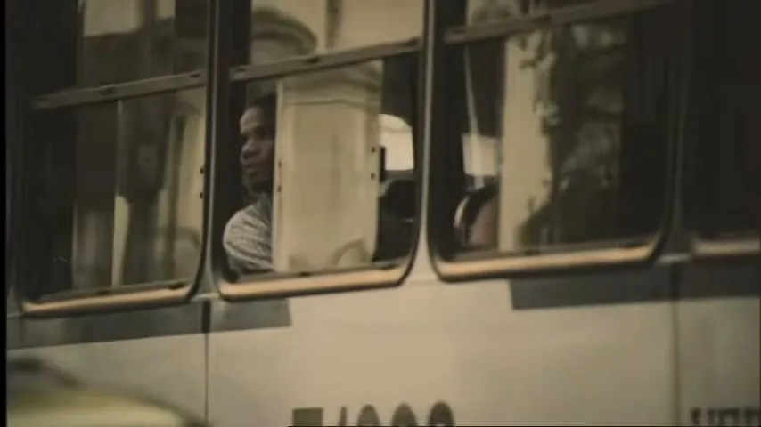 A man with dark skin and a plaid shirt looks out the window of a moving bus or tram, with reflections visible in the glass.