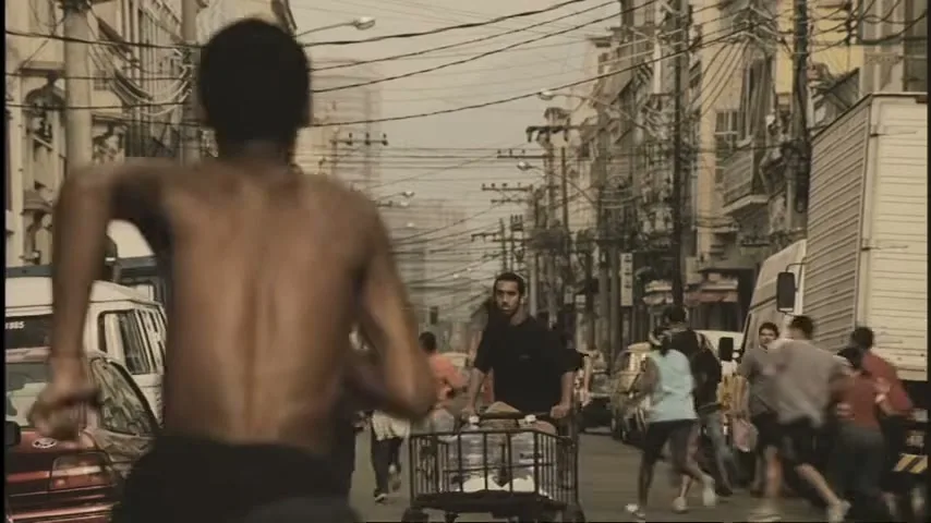 A shirtless young man runs away from the camera down a busy, narrow street filled with people, cars, and tangled power lines.
