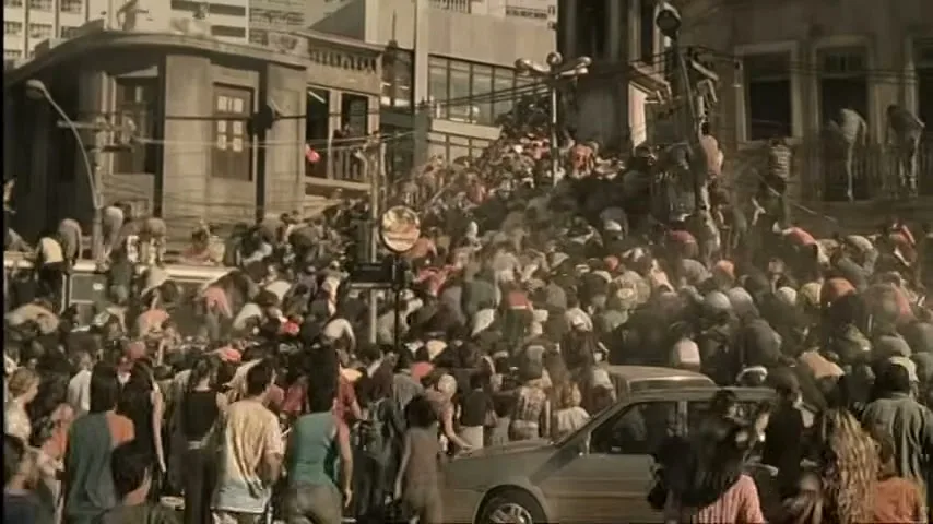 A massive crowd of people, dressed in casual clothes, are seen climbing over cars and buildings in a chaotic urban environment during the day.