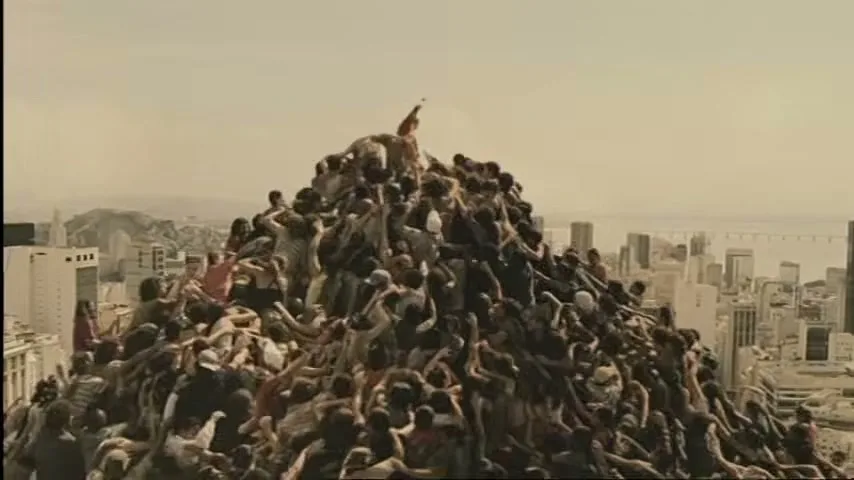 A crowd of people in various casual clothes climb over each other to reach the top of a human mountain, with a city skyline in the background.