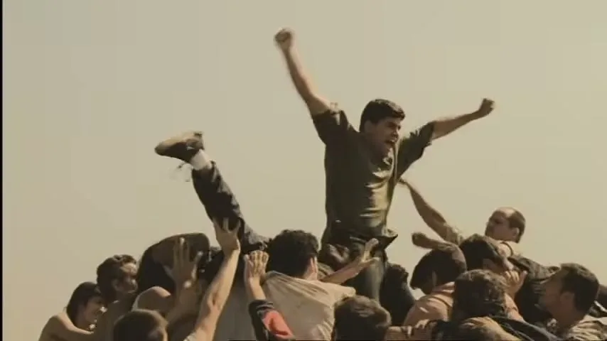 A large group of people are climbing on top of each other, forming a human pyramid, with one man at the top raising his arms in triumph against a clear sky.