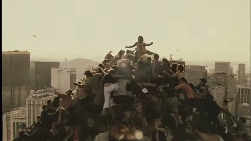 A crowd of people, dressed in casual clothes, are climbing and struggling on a human mountain on a rooftop in a city during the day.