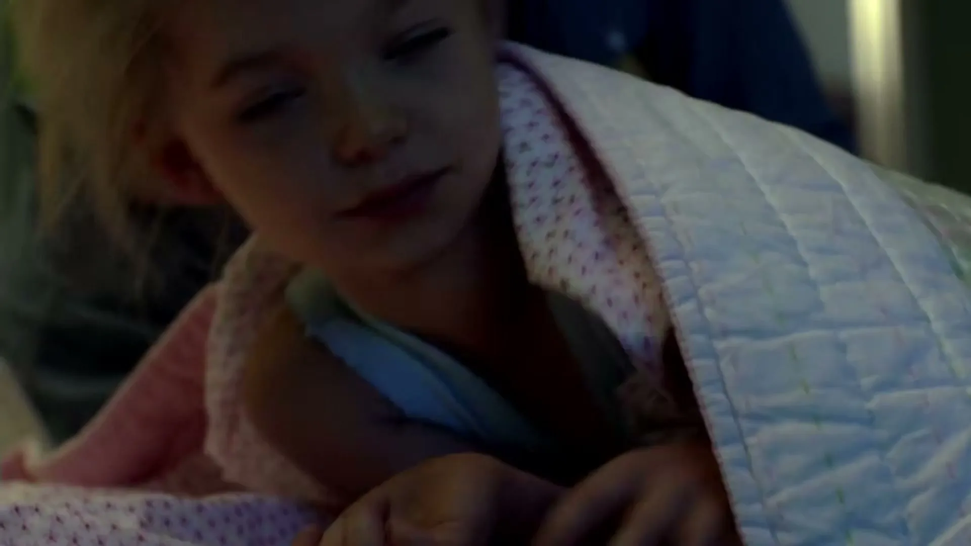 A mother gently wakes her young daughter, who is wrapped in a blanket, in a dimly lit bedroom.