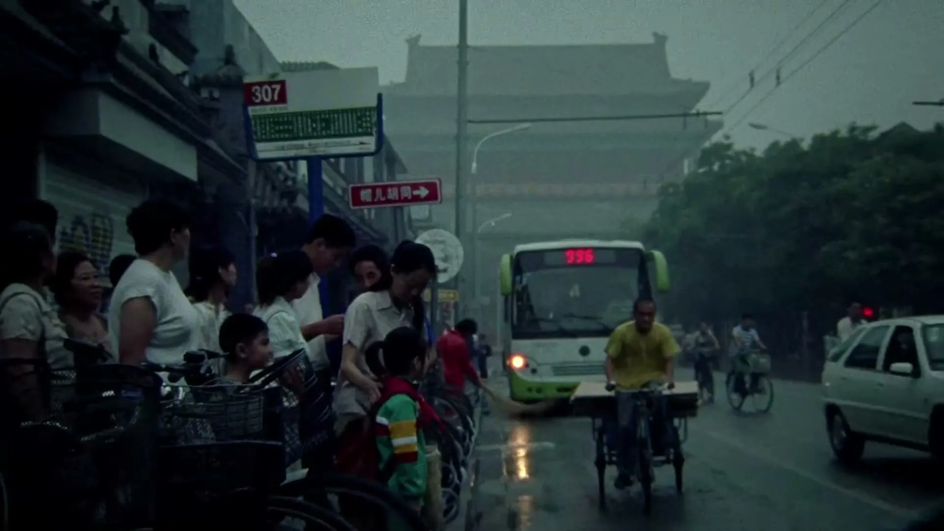 A crowd of people, including children, stand on a wet street next to parked bicycles, while a man on a bicycle rides past in the foreground and a bus approaches in the background on a rainy day.