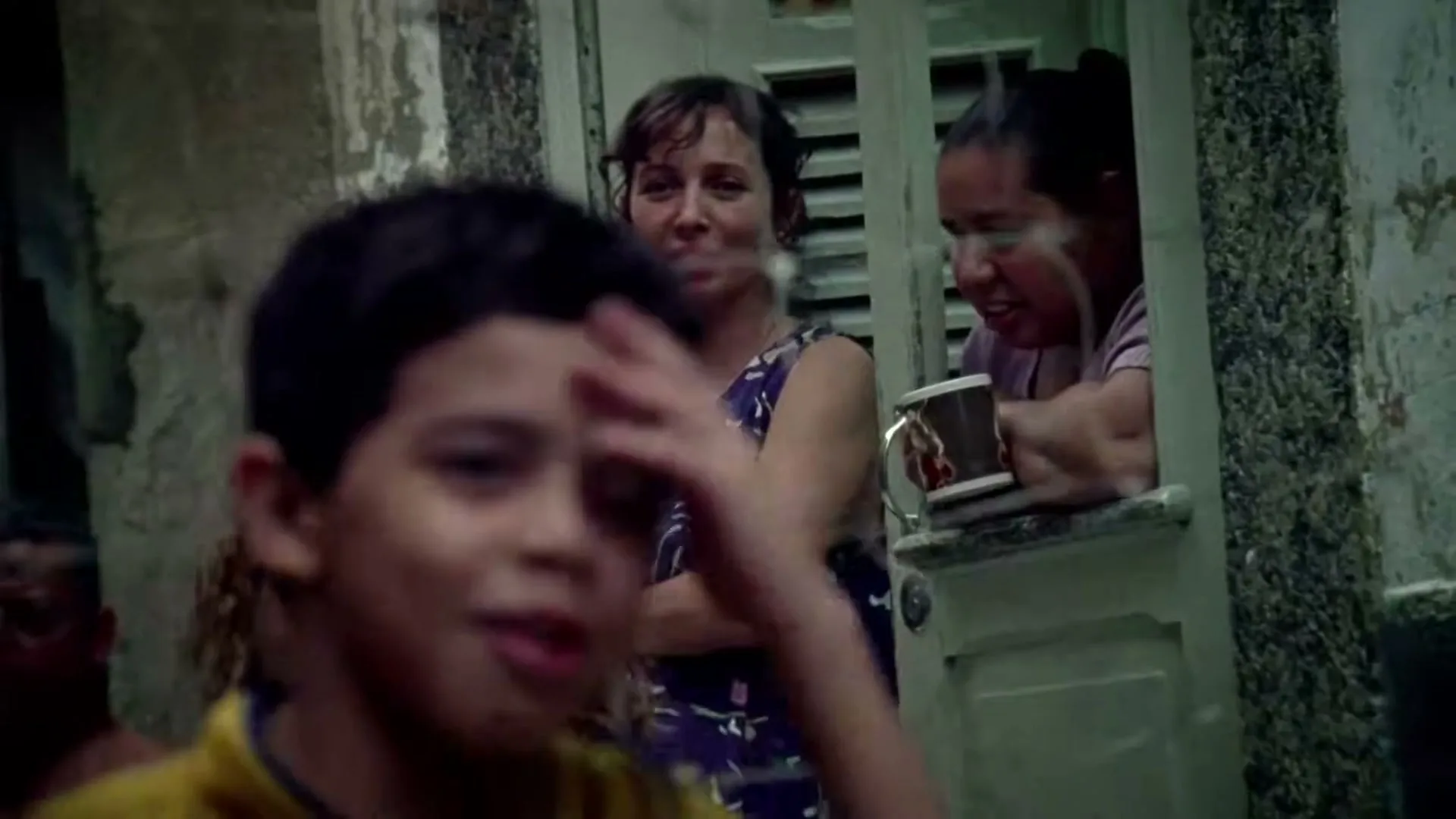 A young boy in a yellow shirt plays in the foreground, while two women watch from a window in a weathered building.