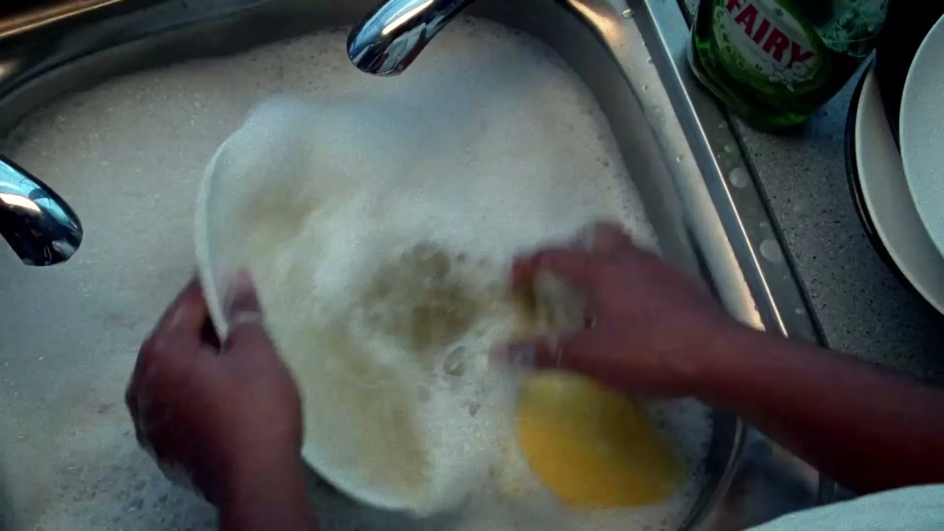A person's hands are seen washing dishes in a bubbly kitchen sink, with a bottle of dish soap visible in the background.