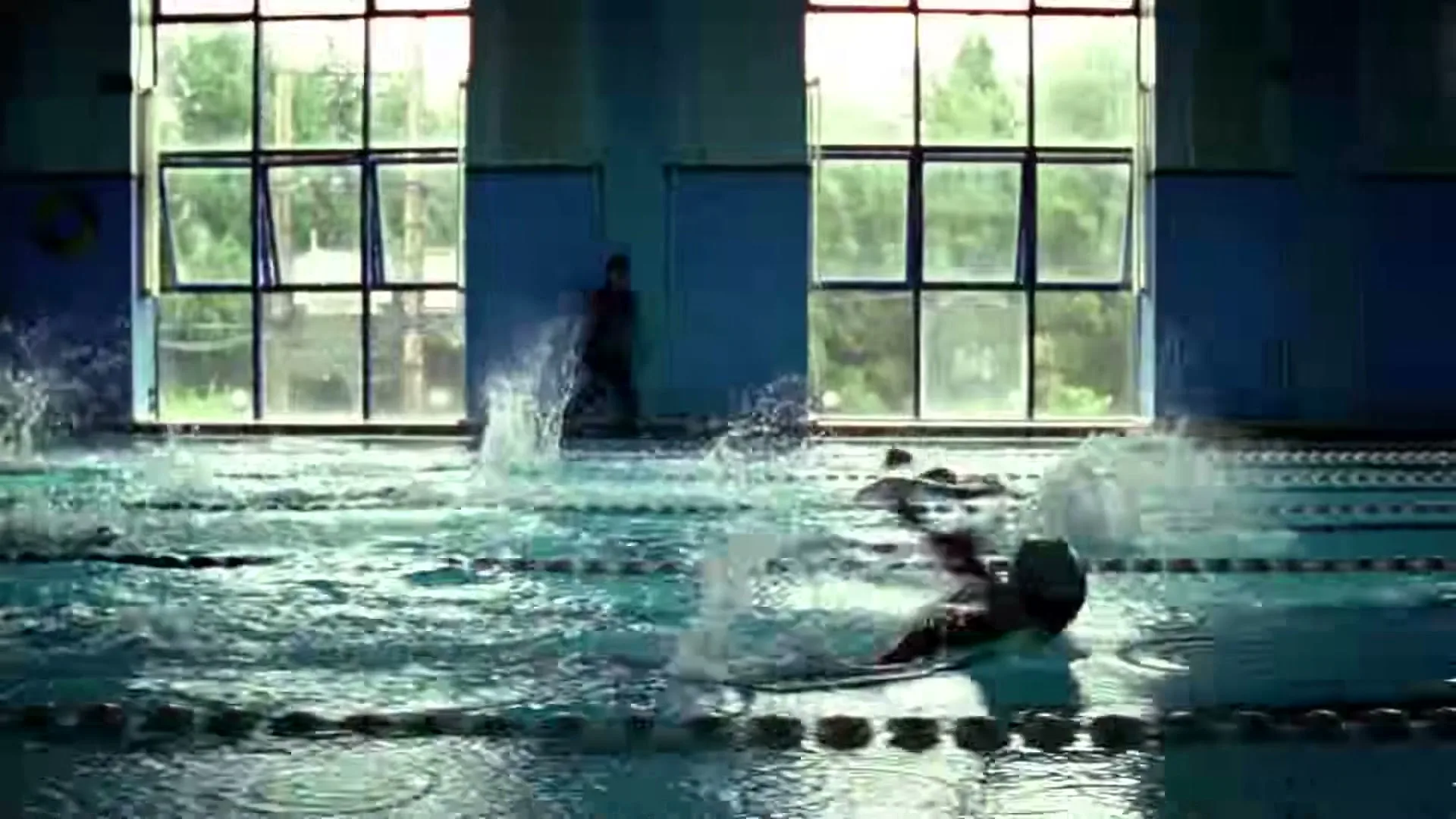 Swimmers create splashes in an indoor pool with large windows in the background, as a person stands by the edge.