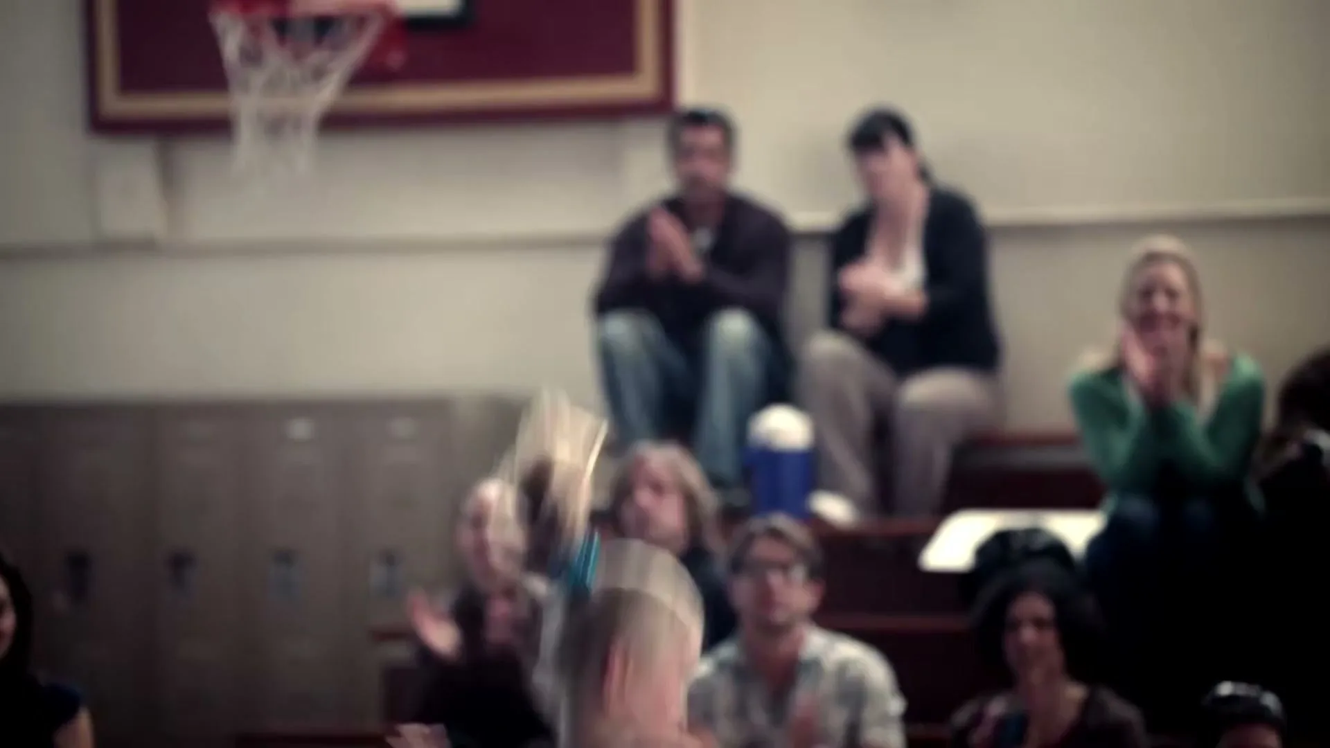 A young blonde girl in a teal leotard performs a gymnastics routine on a balance beam in a school gymnasium, with spectators watching and clapping in the background.