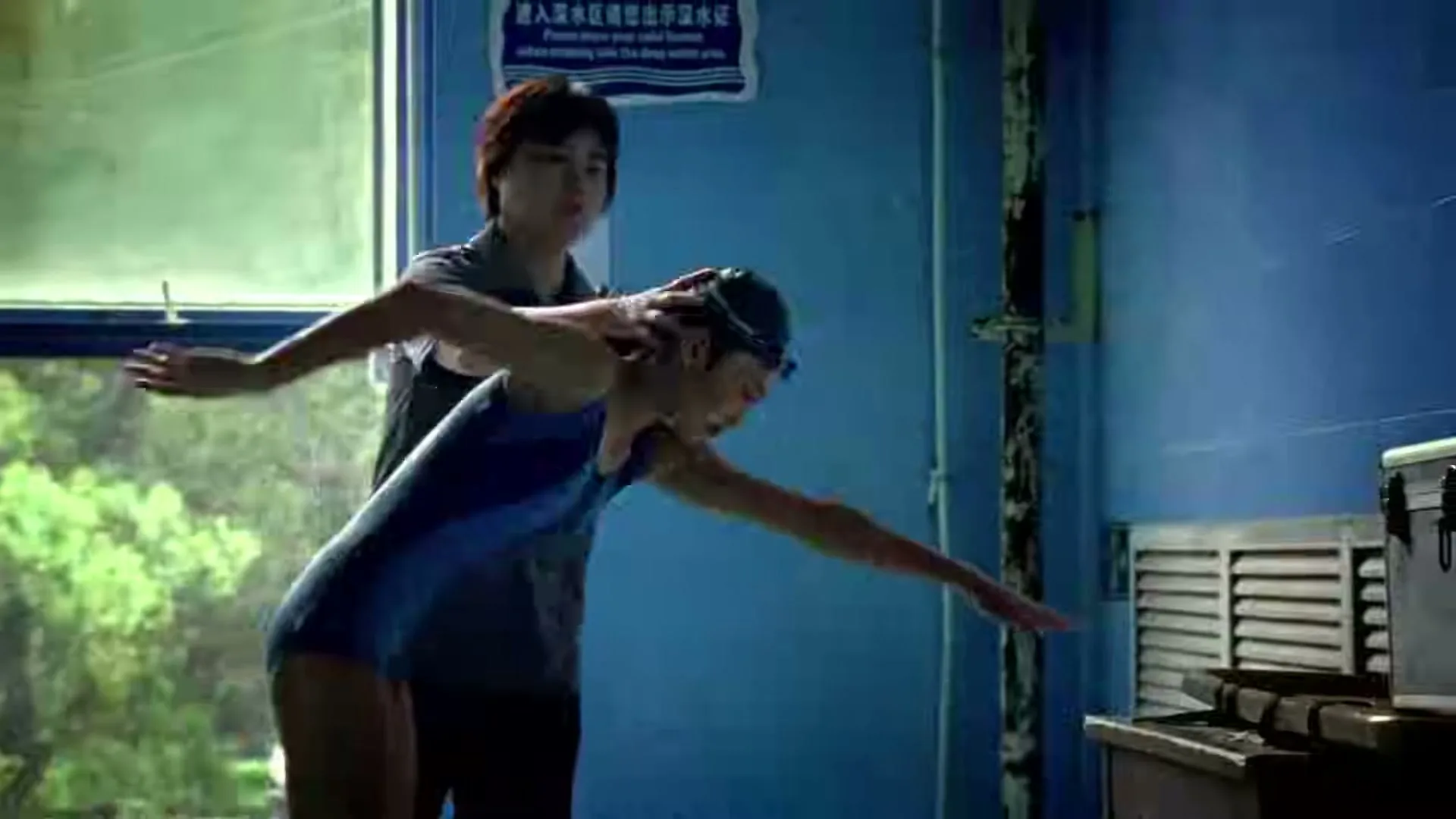 A female coach with short hair helps a young female swimmer practice her dive form in an indoor swimming pool area.