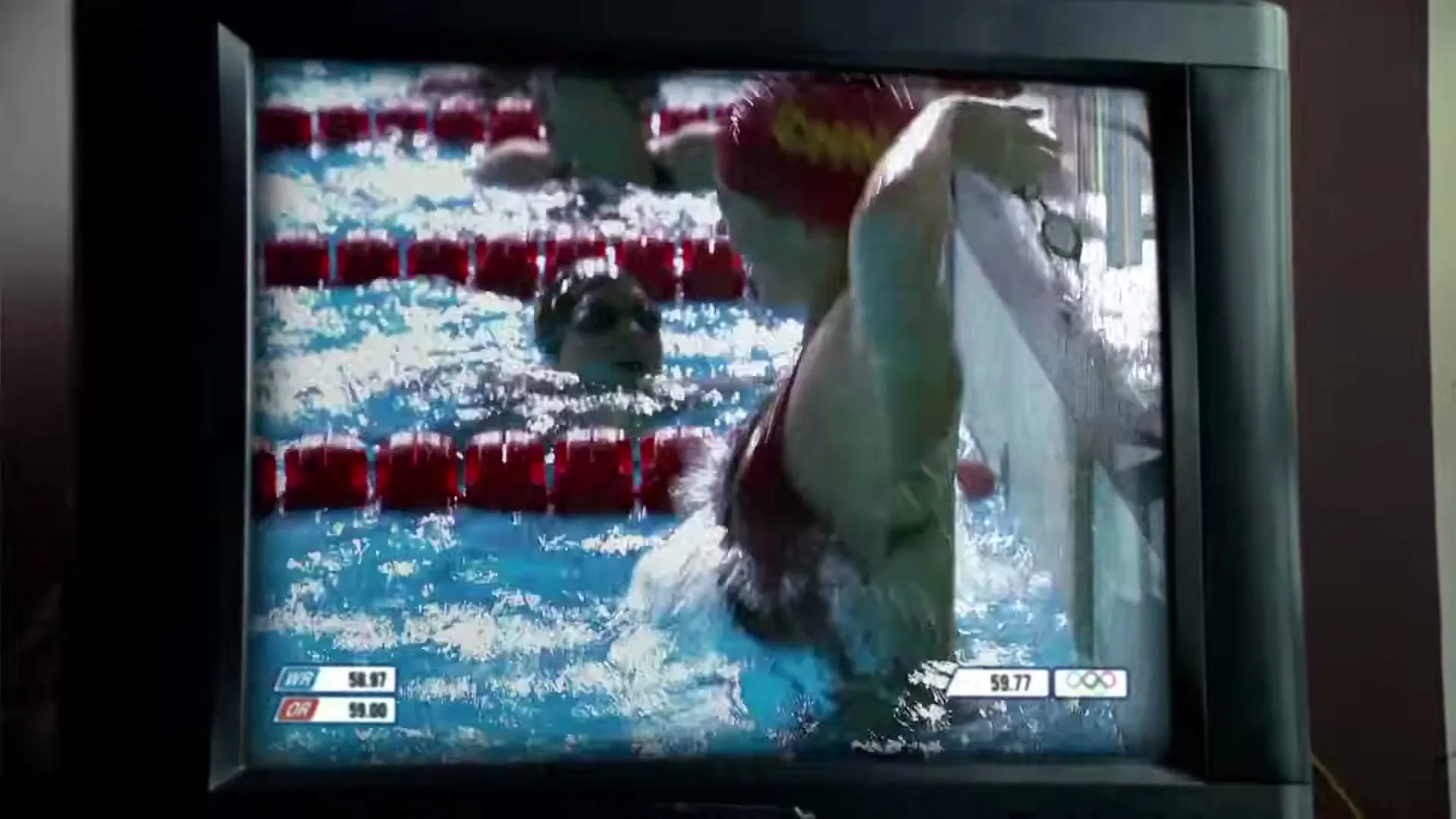 A female swimmer in a red cap with "CHN" on it, emerges from the water in a swimming pool, shown on an old CRT television.