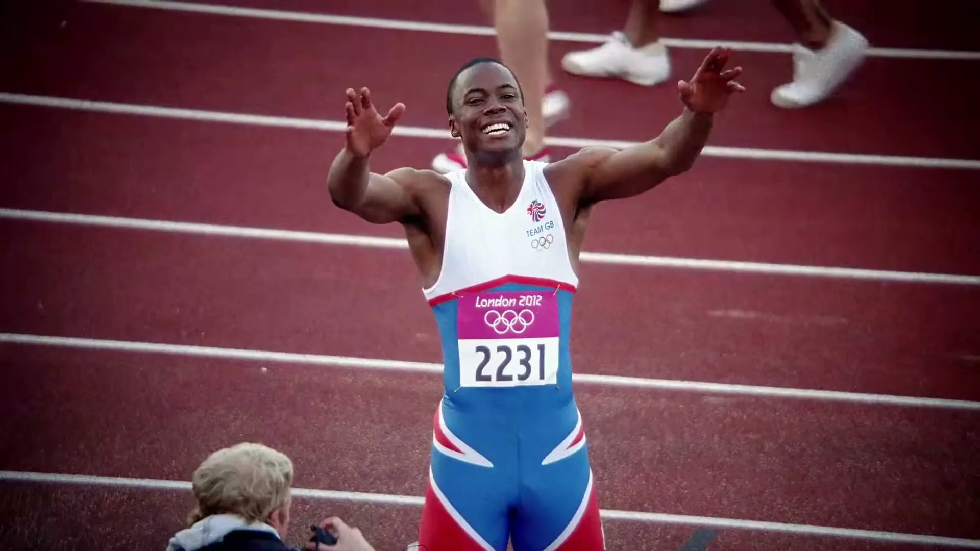 A male athlete in a Team GB uniform celebrates with arms raised on a red running track, with photographers in the foreground.