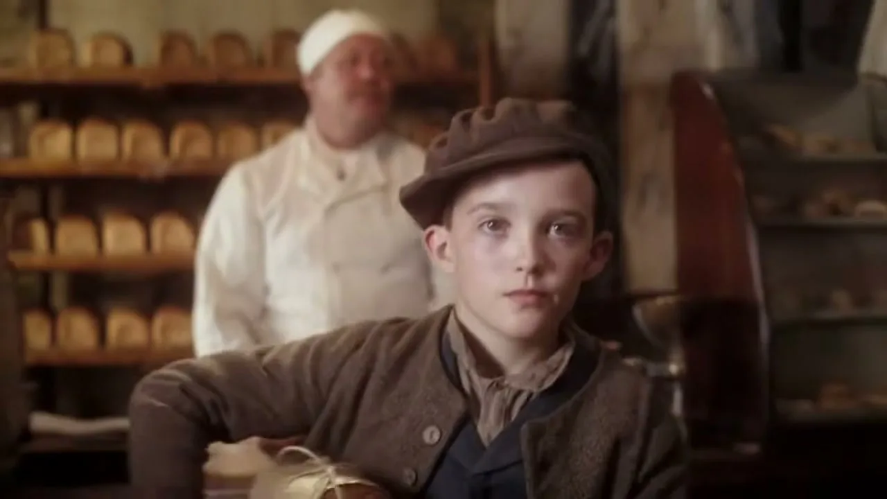 A young boy in a flat cap turns to face the camera, holding a wrapped loaf of bread in a rustic bakery with shelves of bread behind him.