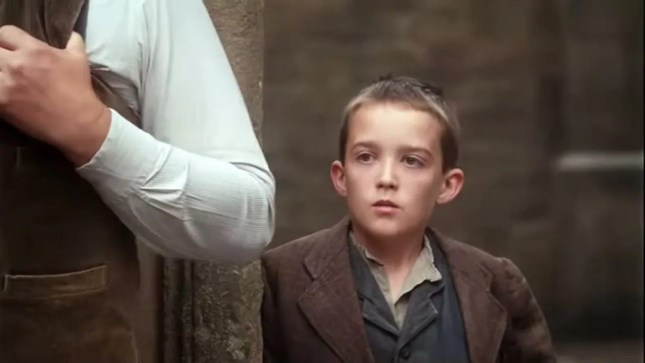 A young boy with a brown jacket and a light shirt looks up with a curious expression, standing next to an adult in a vest.