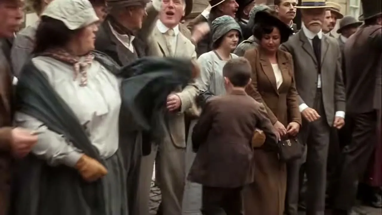 A crowd of people in early 20th-century attire watch a boy run past them on a street.