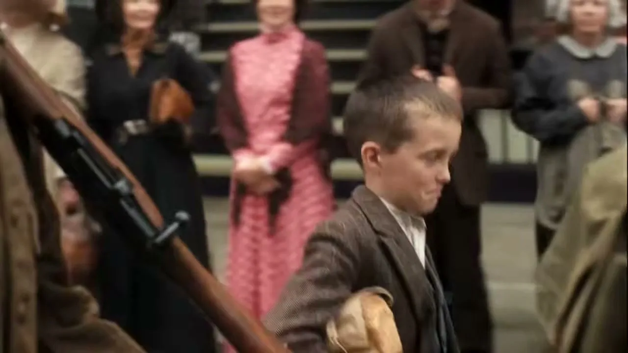 A young boy in period clothing, holding a loaf of bread, looks around nervously as he walks past a rifle and other people.