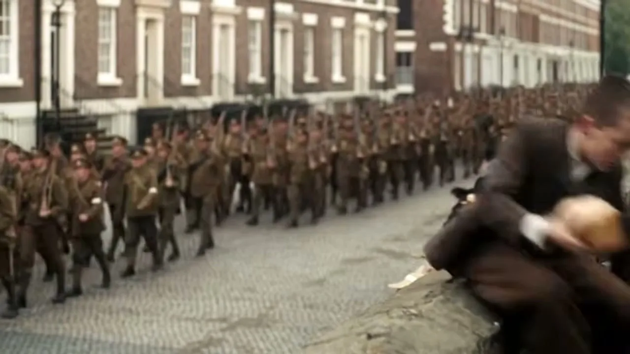 A young boy in old-fashioned clothes sits on a stone wall, holding a loaf of bread, as soldiers march past on a cobbled street.