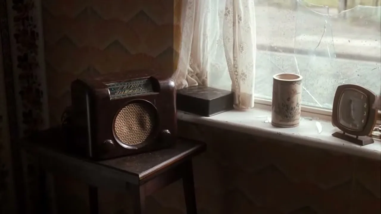 A vintage radio sits on a small table next to a window with a cracked pane, as a child looks in from outside.