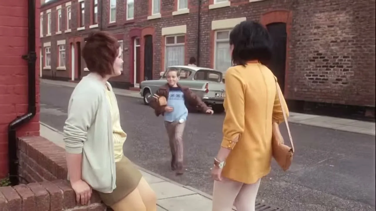 A young boy runs down a street towards two women, carrying a loaf of bread under his arm in a working-class neighborhood.