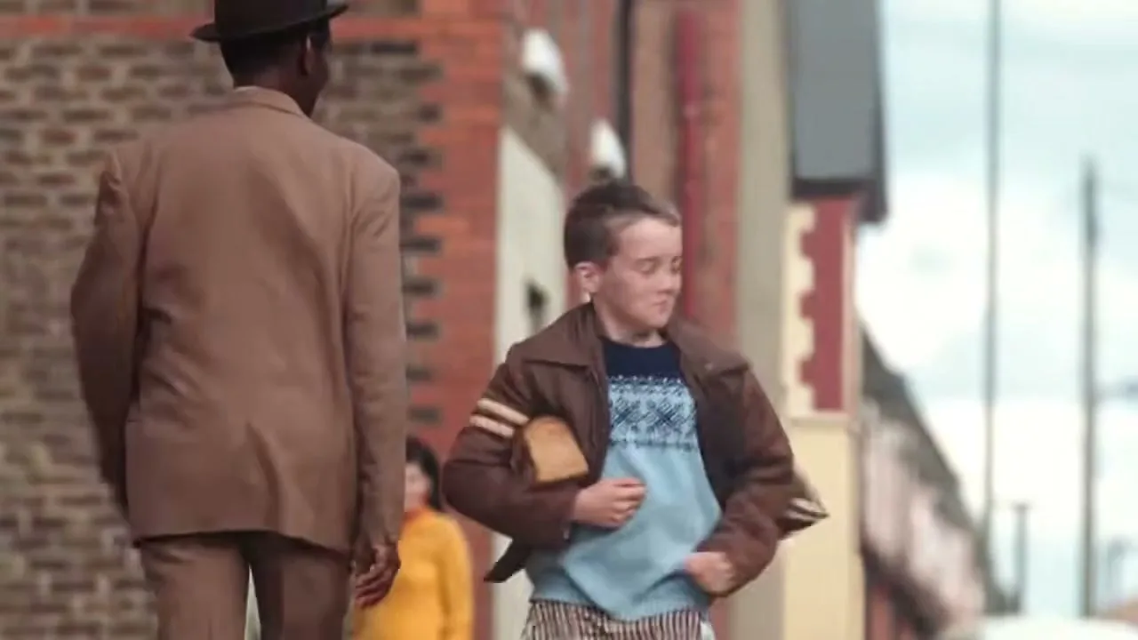 A young boy in a brown jacket runs past a man in a suit, holding a slice of bread, on a street in a working-class neighborhood.