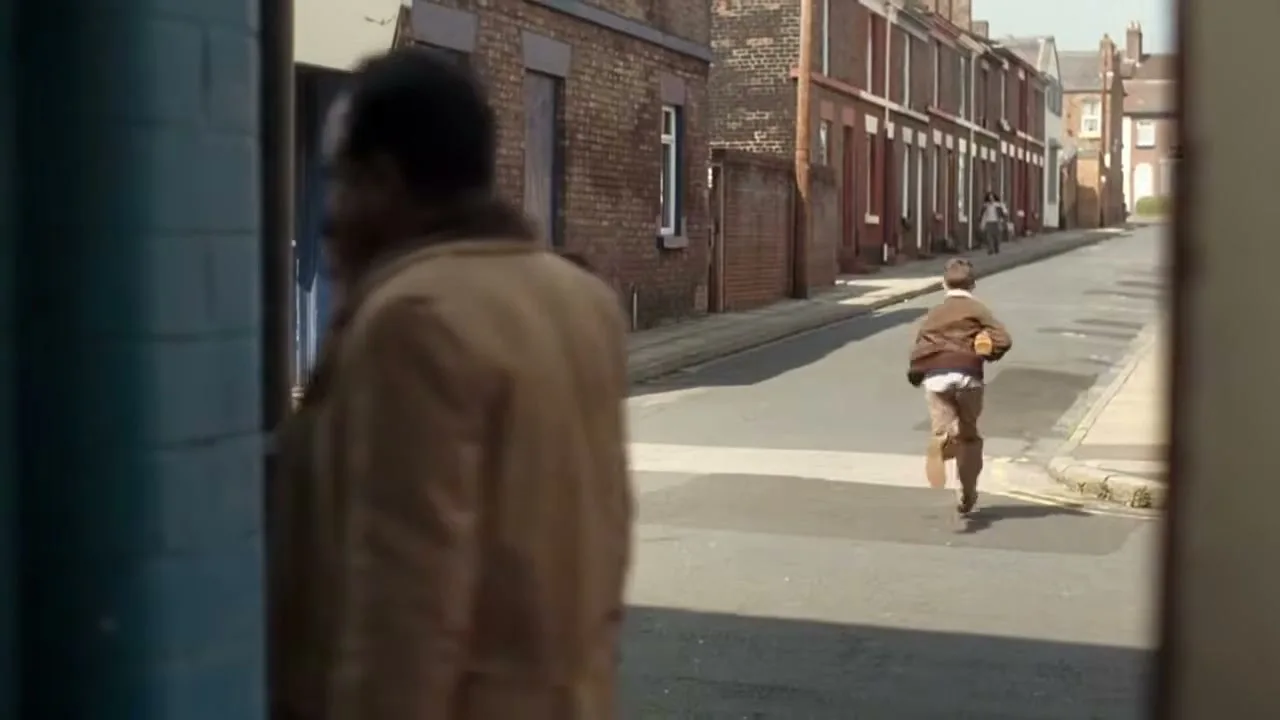 A young boy in a brown jacket and shorts runs down a cobbled street in a working-class neighborhood, carrying a loaf of bread.