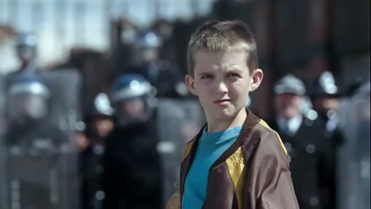 A young boy in a brown and yellow jacket stands in front of a line of riot police, looking determined.
