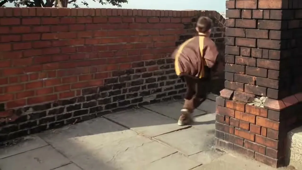 A young boy in a brown and yellow jacket runs past a brick wall and pillar on a sunny day.