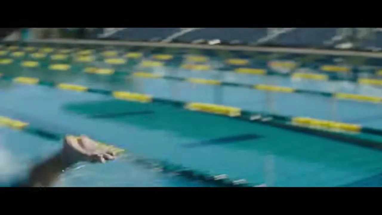 A swimmer's foot pushes off a starting block, then plunges into the water, creating a splash in a bright outdoor pool.