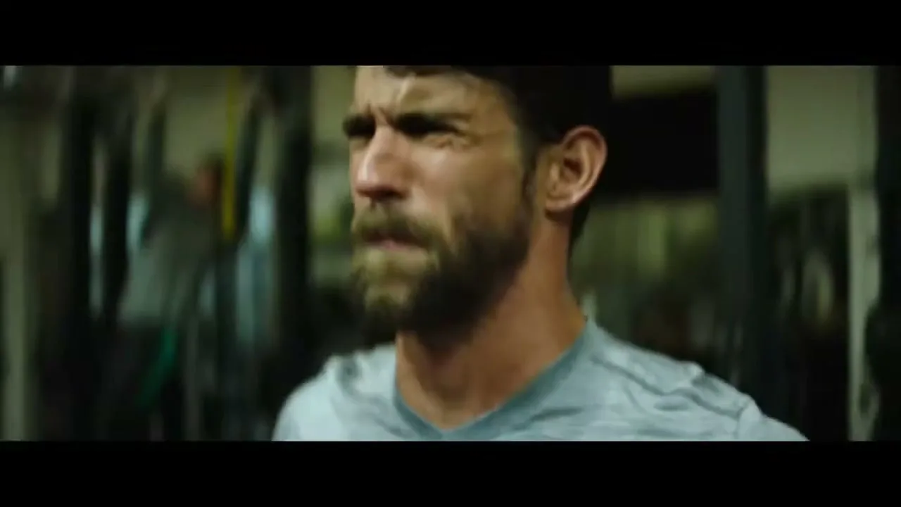 A bearded man in his 30s, wearing a grey t-shirt, runs on a treadmill with a determined expression in a dimly lit gym.