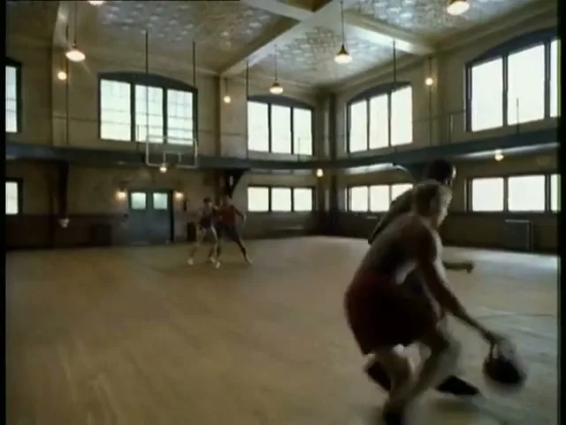 A basketball player in a yellow jersey dribbles the ball on a court, while another player in a red jersey defends him in a wide shot.