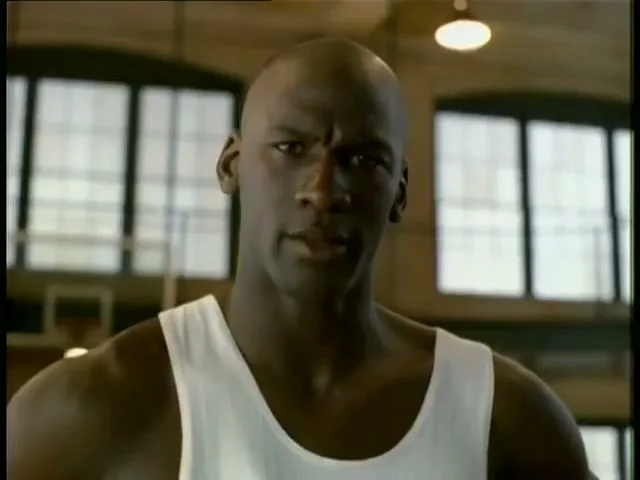 Michael Jordan, a bald Black man in a white tank top, looks directly at the camera in a basketball gym.