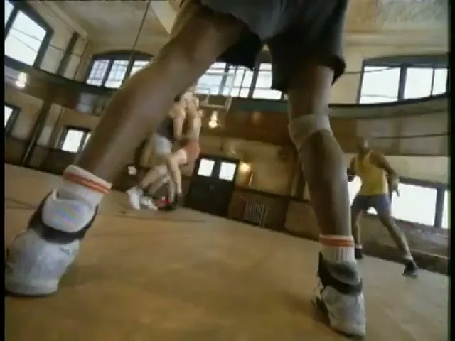 A low-angle shot shows a basketball player's legs in sneakers and shorts, with an animated red character in the foreground, on a wooden court.