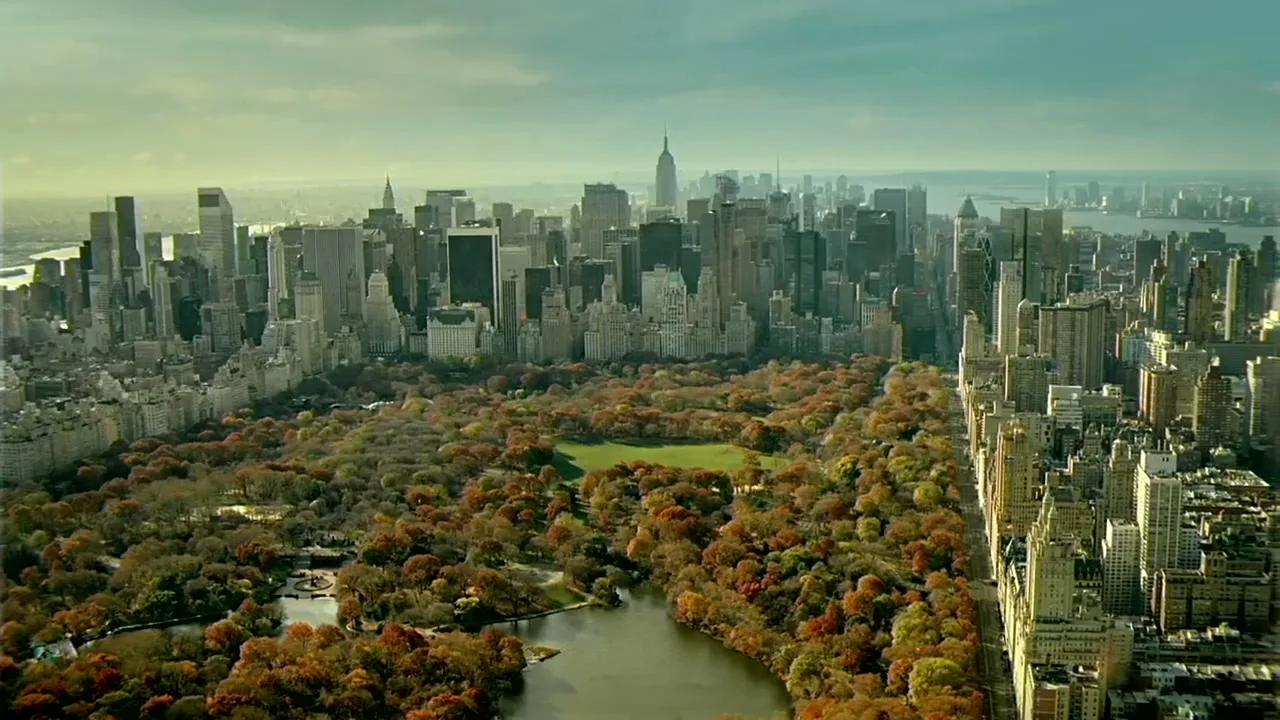 An aerial view of Central Park in New York City, surrounded by skyscrapers, with autumn trees and a lake visible under a bright sky.