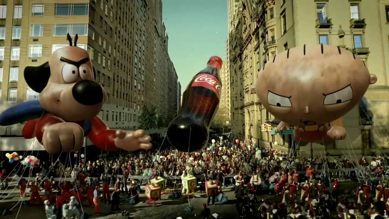 Two large cartoon character balloons and a Coca-Cola bottle balloon float above a crowd of people during a parade on a city street.