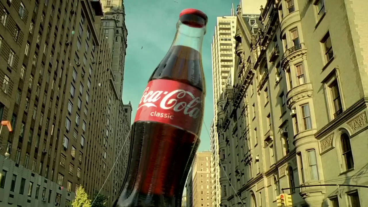 A giant Coca-Cola Classic bottle stands tall between two buildings on a city street, with a clear blue sky above.