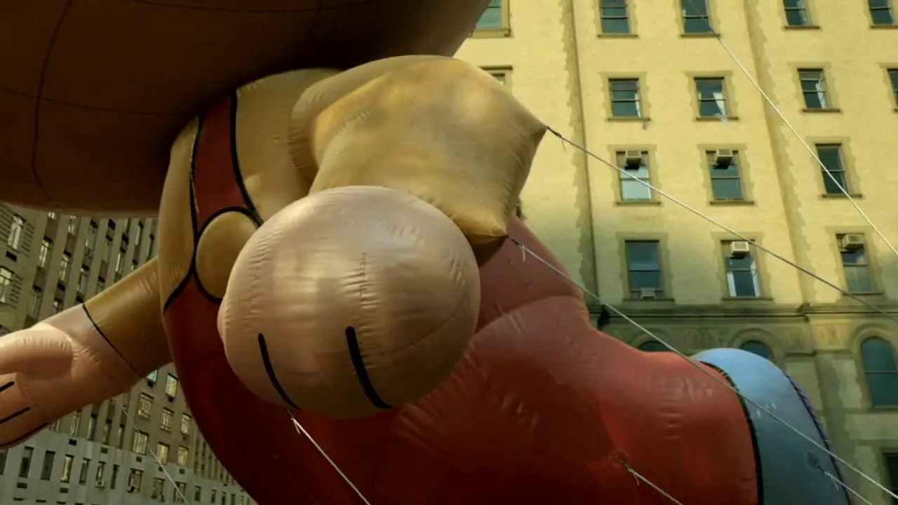 A large, inflatable cartoon character balloon, red and tan, floats past a tall, light-brown building with many windows during a parade.