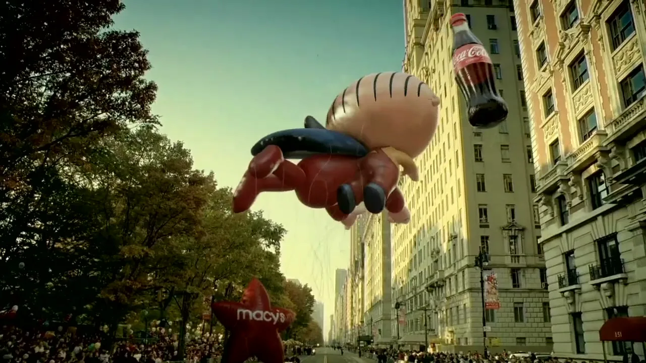 A giant Stewie Griffin balloon from Family Guy, holding a Coca-Cola bottle, floats above a Macy's star balloon during a parade on a city street.