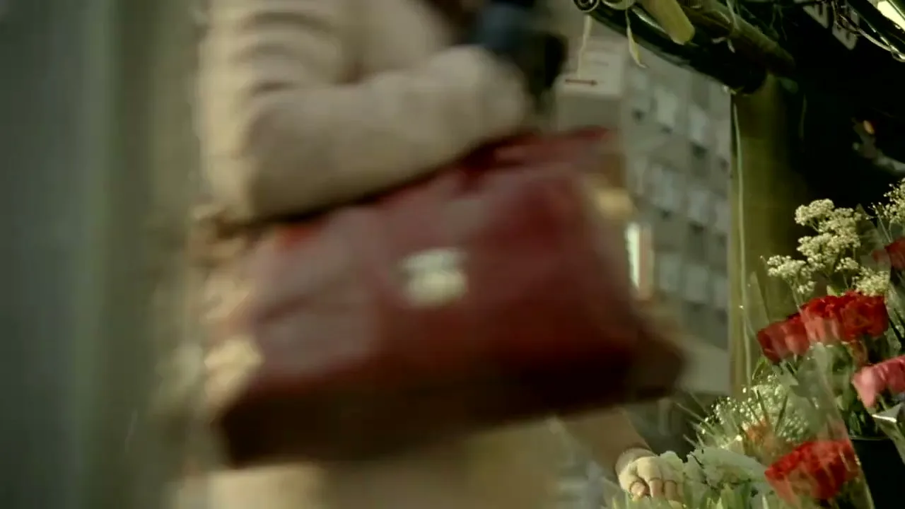 A male flower vendor in a brown cap and vest arranges flowers at his street stall in a city, looking up as a woman passes by.