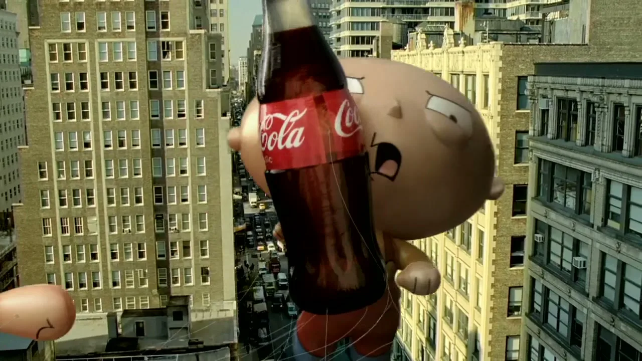 A giant Stewie Griffin balloon from Family Guy holds a Coca-Cola bottle while floating above a city street with tall buildings.