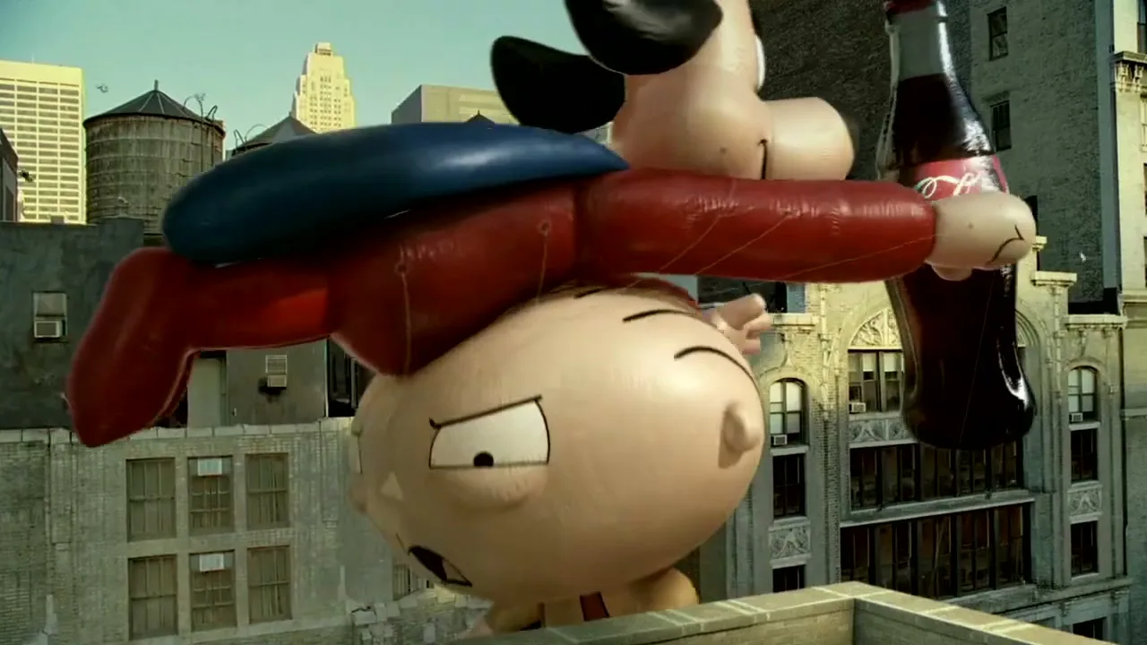 An inflatable Stewie Griffin from Family Guy is seen from a high angle, struggling to hold onto a giant Coca-Cola bottle in a city.