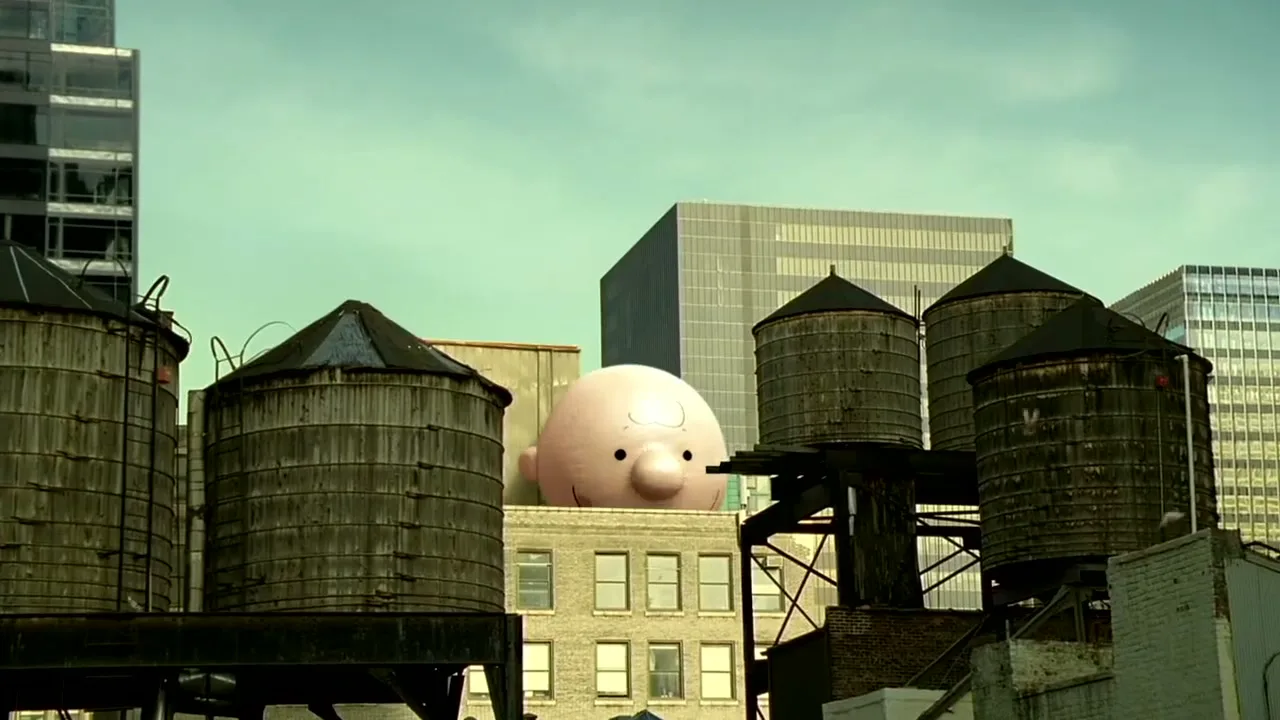 A giant Charlie Brown balloon peeks over a building, surrounded by water towers on a city rooftop during the day.