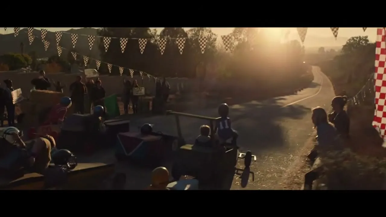 A wide shot of a soapbox derby race with children in various homemade cars at the starting line, with spectators and checkered flags under a golden hour sky.