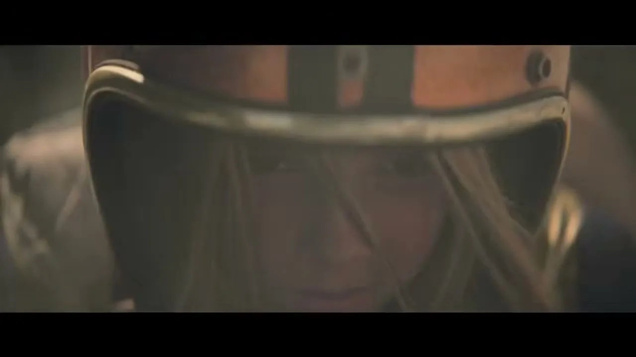 A young girl with blonde hair wearing an orange racing helmet looks intently forward, as if preparing for a race.