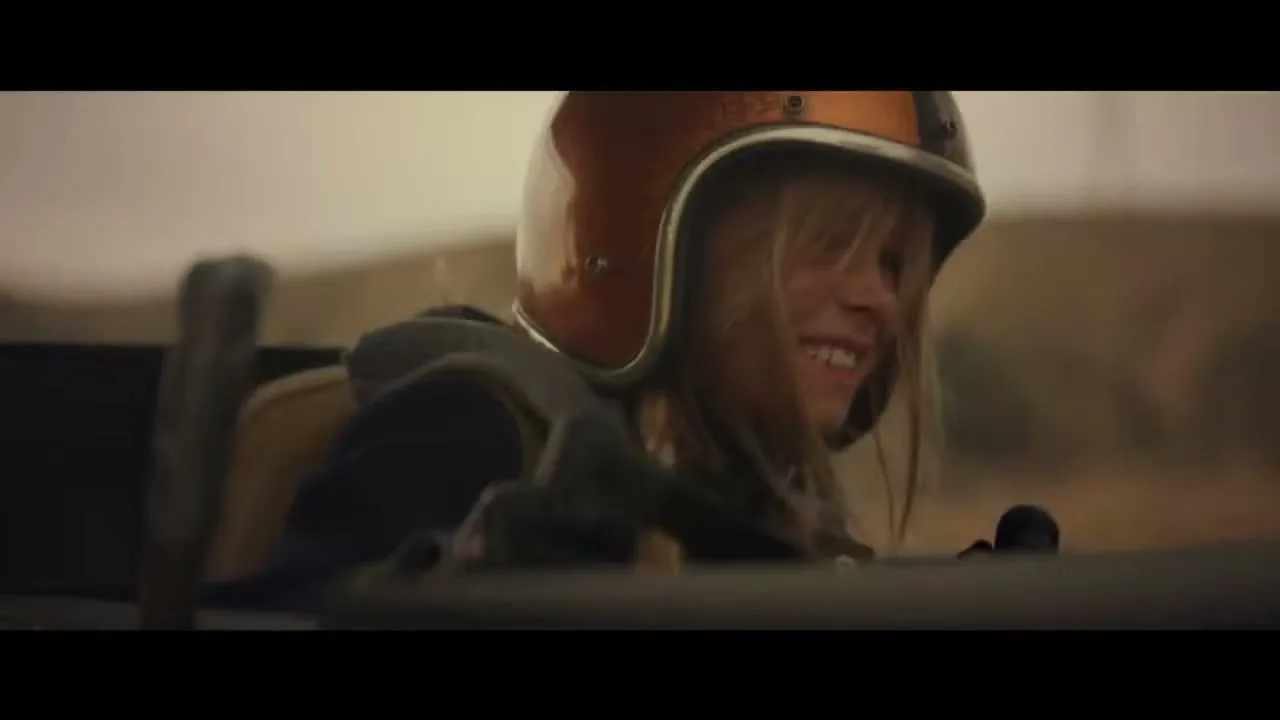 A young girl with blonde hair and a racing helmet smiles excitedly while driving a car in a desert landscape.